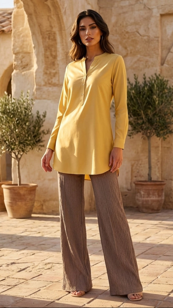Woman in a yellow top and brown pants standing in a sunlit outdoor setting with arches and palm trees.