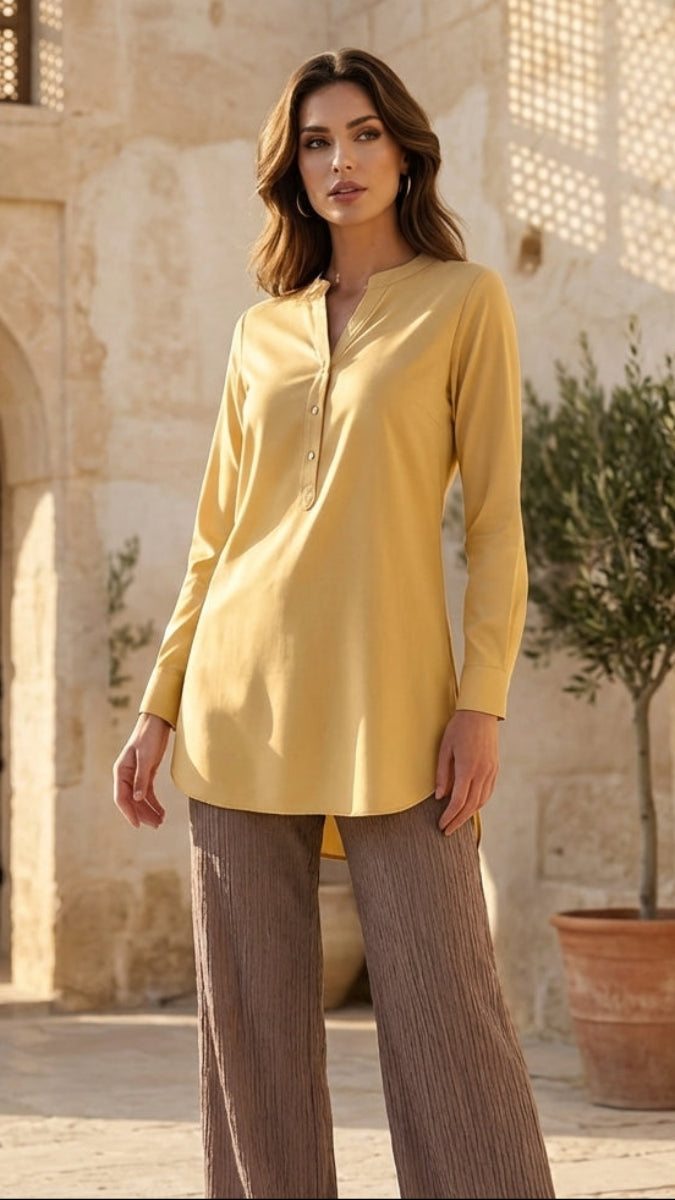Woman in a yellow shirt and brown pants standing in front of a stone building with a tree in the background.