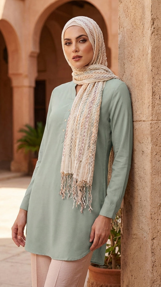 Woman wearing a light green dress and beige hijab in an outdoor setting with arches and plants.