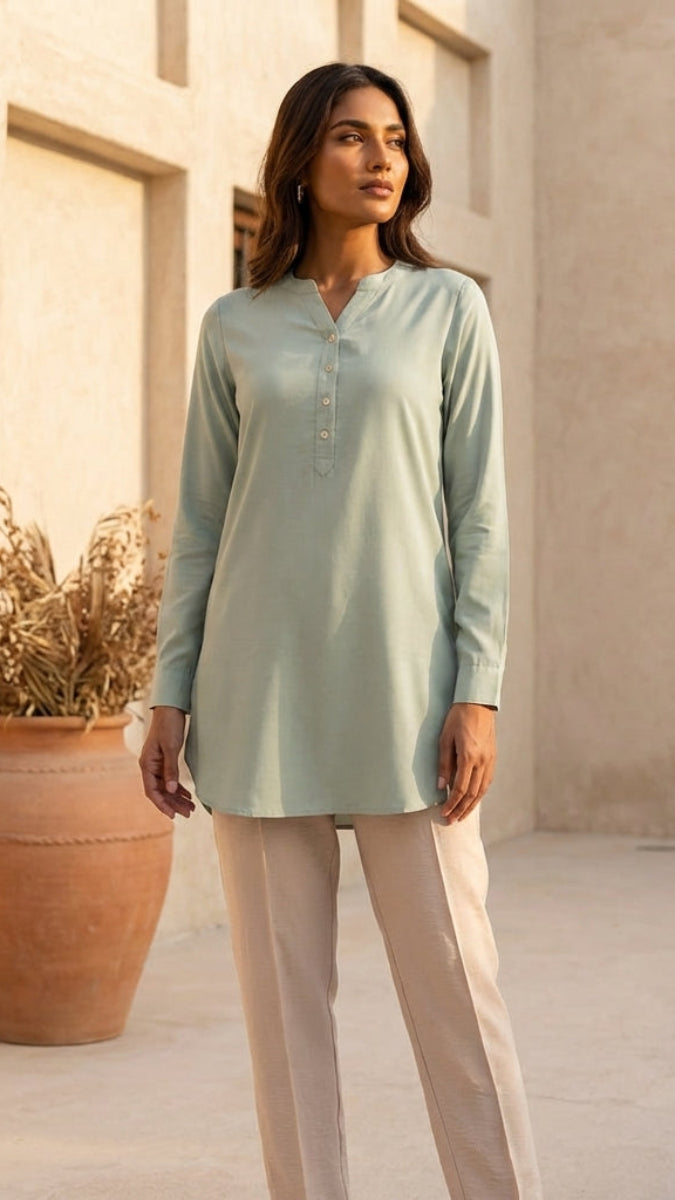 Woman in a light green long-sleeve top and beige pants standing in front of a textured wall with a pot of dried plants.