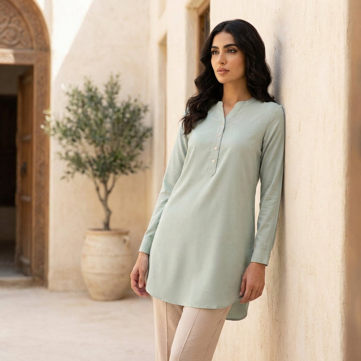 Woman wearing a light green dress leaning against a stone wall with a plant in the background.