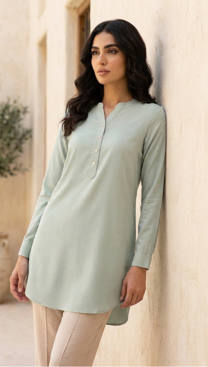 Woman wearing a light green dress leaning against a stone wall with a plant in the background.