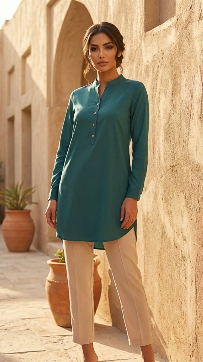 Woman in teal shirt and beige pants standing in a sunlit courtyard with stone walls and potted plants.
