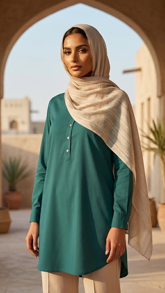 Woman in a teal dress and beige hijab standing in a sunlit outdoor setting with arches and plants.