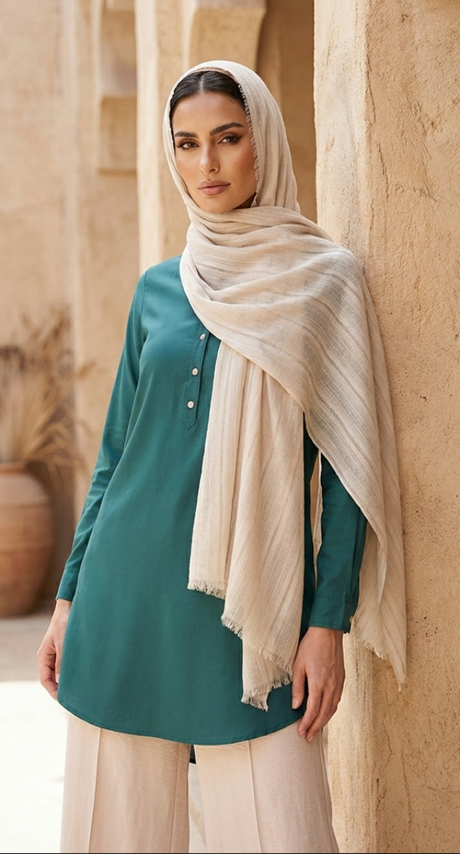 Woman wearing a teal top and beige hijab standing against a beige wall.