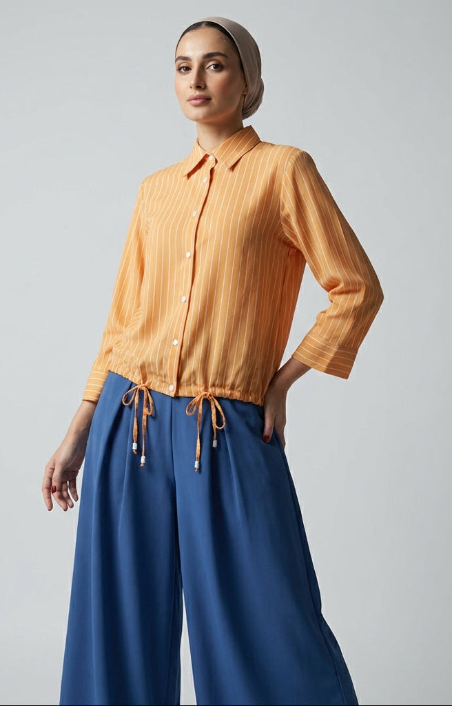 Woman wearing a saffron stripe shirt and blue pants against a gray background