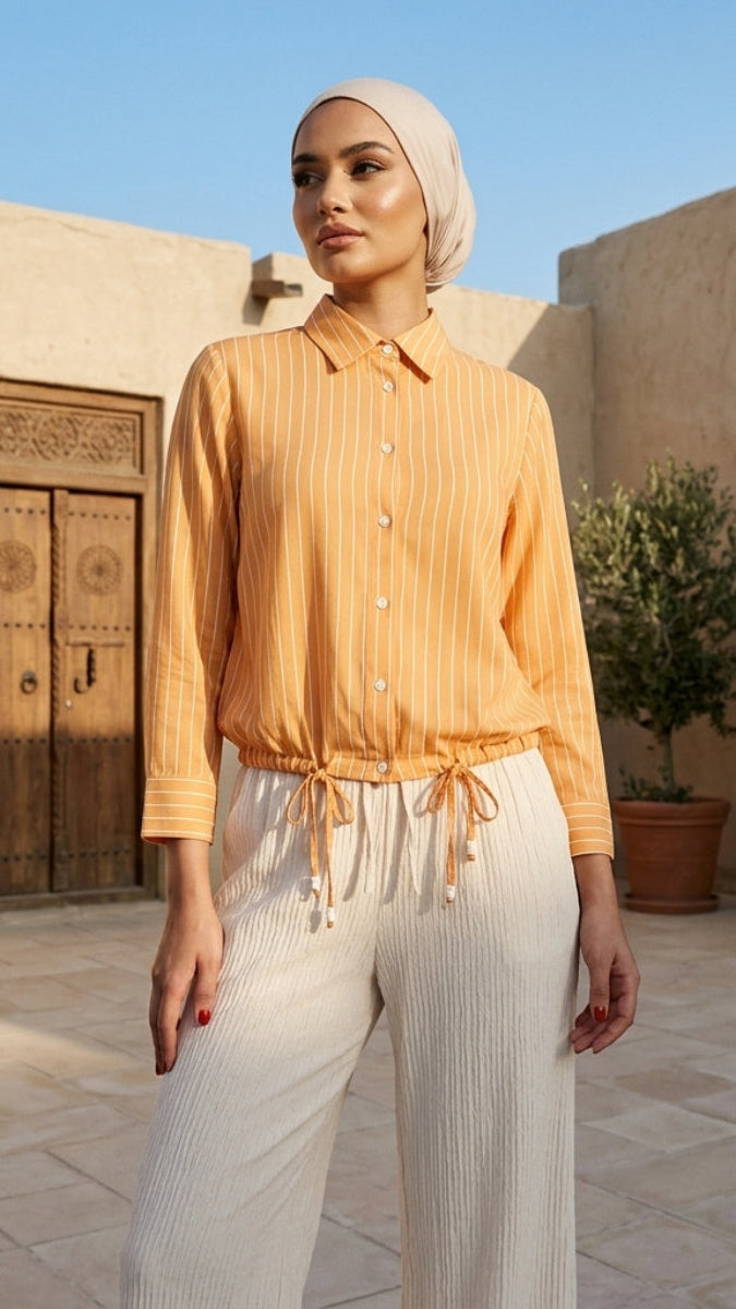 Woman in a mustard yellow blouse and white pants standing in a sunlit courtyard with architectural elements.