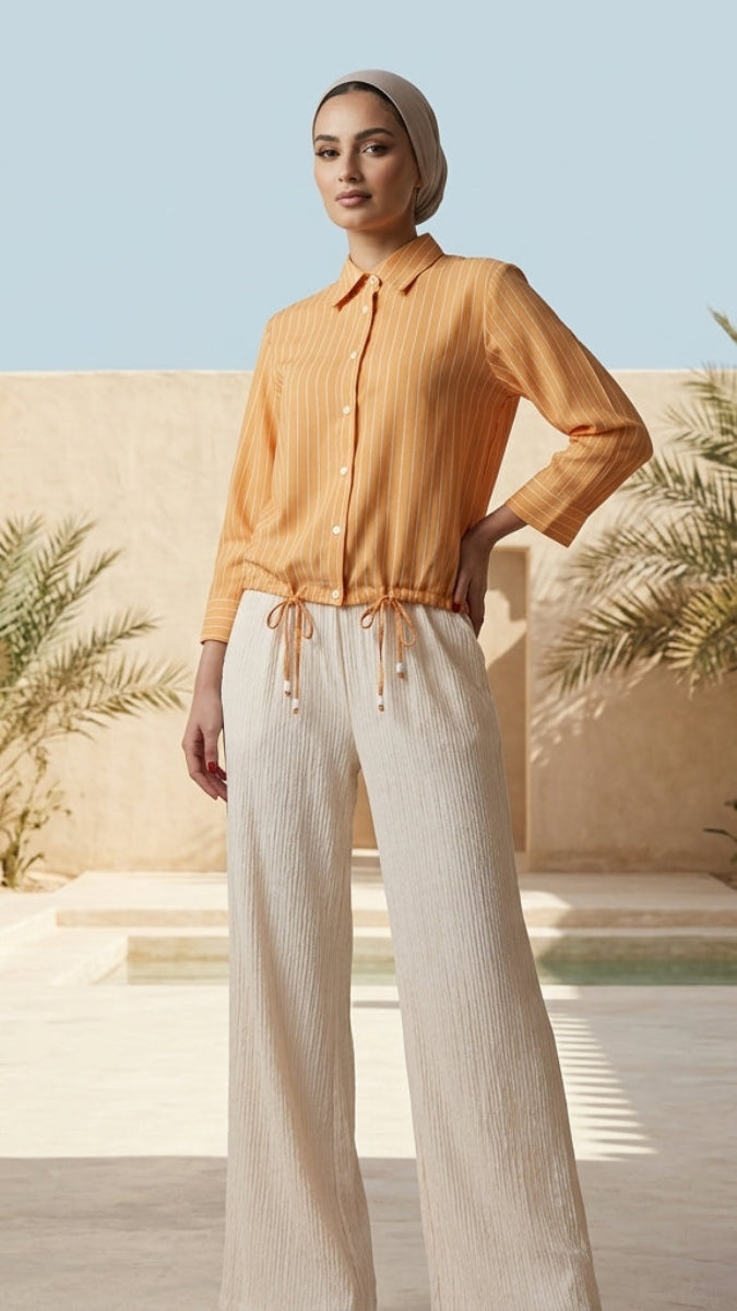 Woman in an orange shirt and white pants standing in a sunlit outdoor setting with plants and a building.