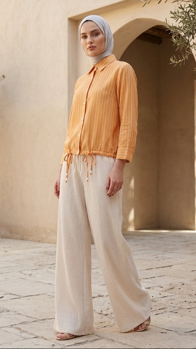 Woman in orange top and white pants standing in a sunlit outdoor setting with potted olive trees.