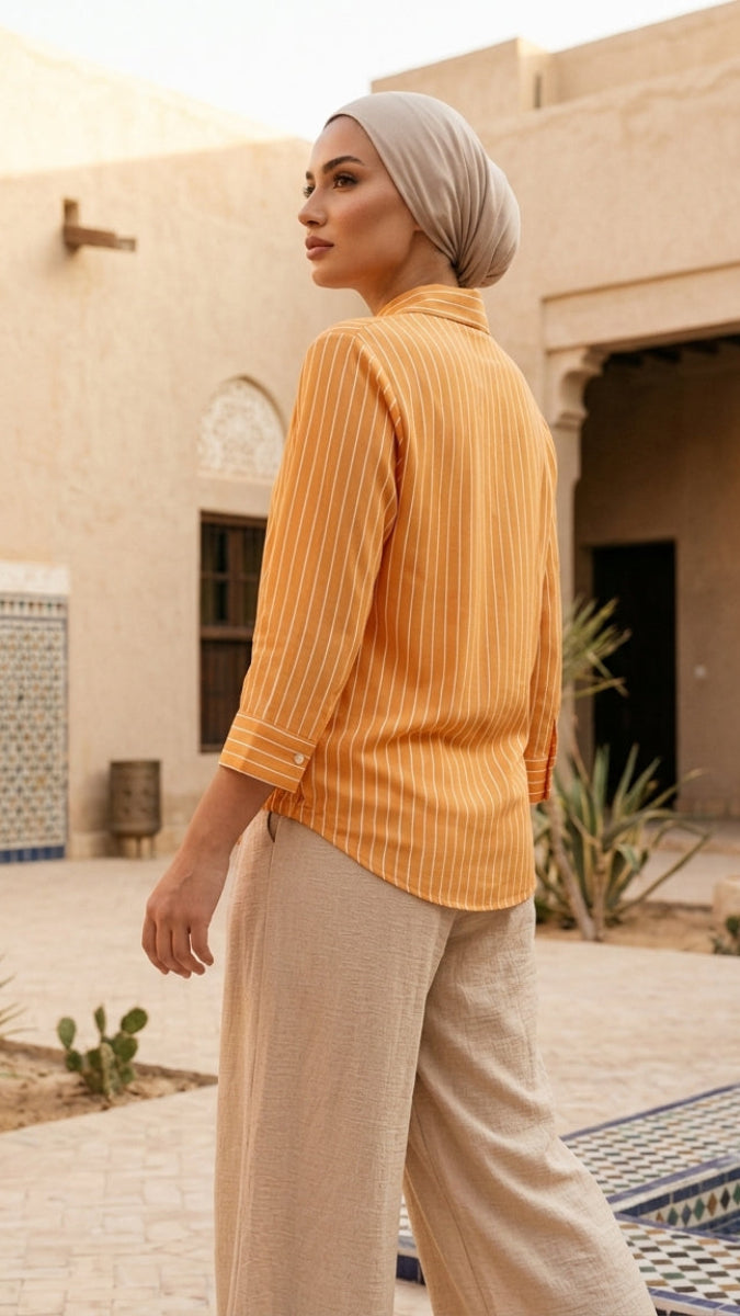 Woman in a yellow top and beige hijab standing in front of a traditional building with arches and plants.