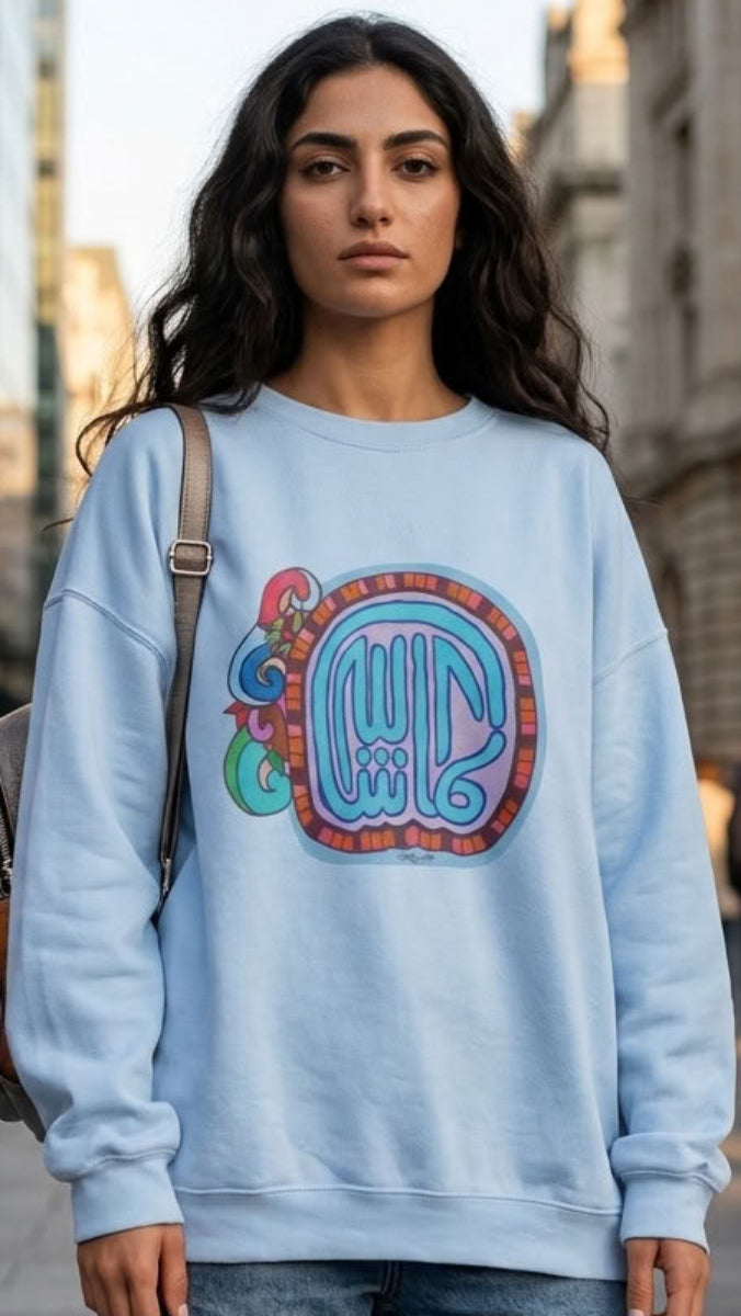 A woman stands on a city street wearing a light blue crewneck sweatshirt with a colorful retro graphic on the front, paired with distressed blue jeans and a brown shoulder bag.