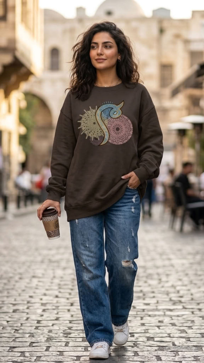 A woman walking down a cobblestone street wearing a dark brown crewneck sweatshirt with a colorful graphic print, blue jeans, and white sneakers, holding a coffee cup.