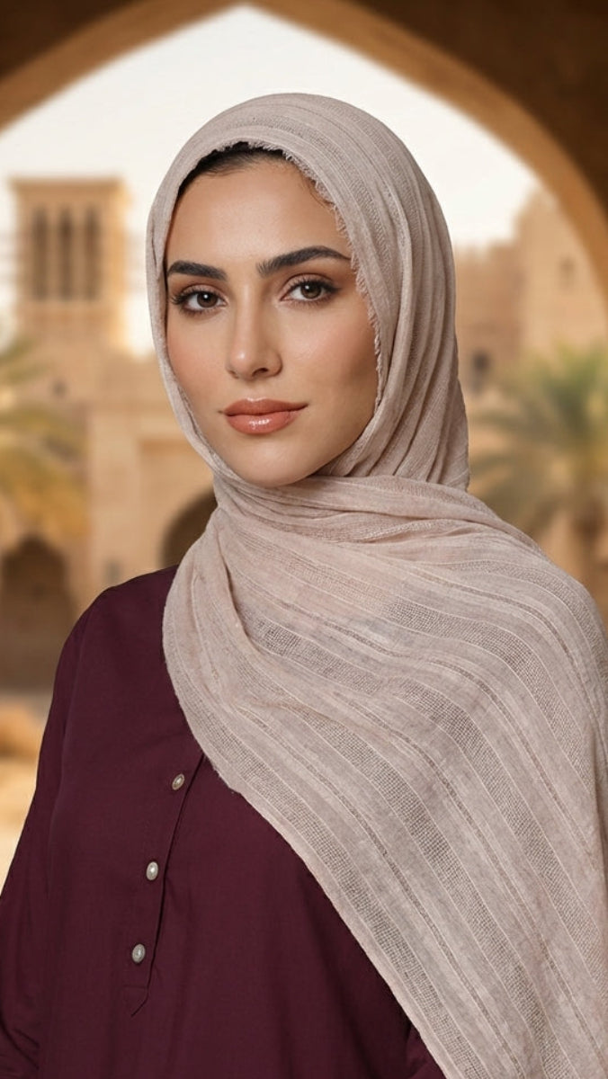 Woman wearing a beige hijab and maroon outfit in an outdoor setting with arches and greenery.