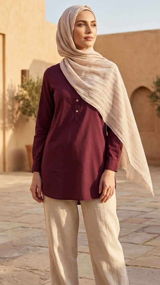 Woman wearing a hijab and maroon top in an outdoor setting with plants and stone pavement.