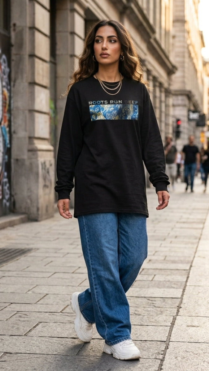 A woman walking on a city street wearing a black long-sleeve graphic t-shirt with a colorful rectangular chest print, blue jeans, and white sneakers.