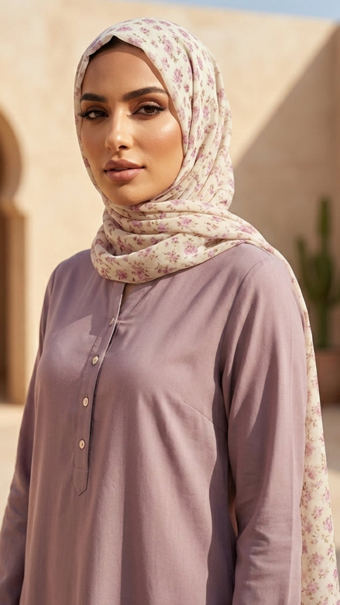 Woman wearing a light purple shirt and floral hijab standing in a desert-like setting with arches and plants.