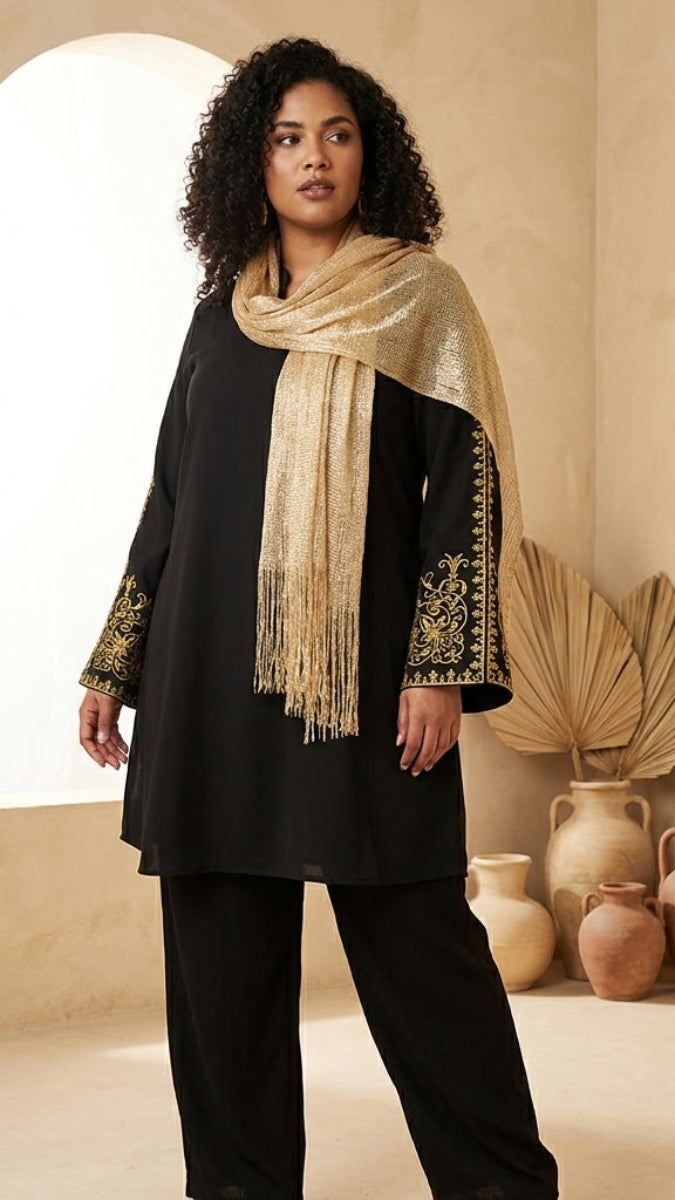 Woman wearing a black outfit with gold embroidery and a beige scarf in a warm-toned room.