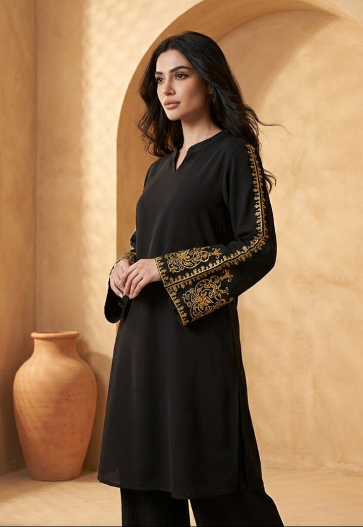 Woman in a black outfit with gold embroidery standing in a warm-toned interior setting.