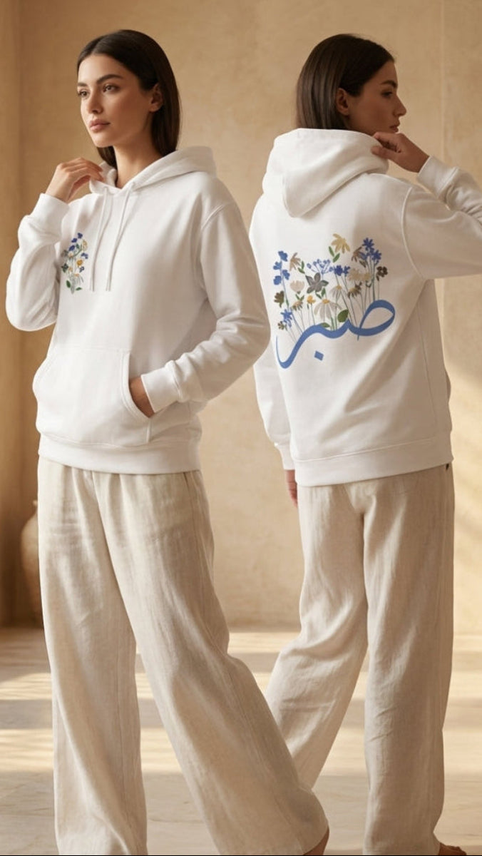 Two women wearing white hoodies with floral designs in a sunlit room.