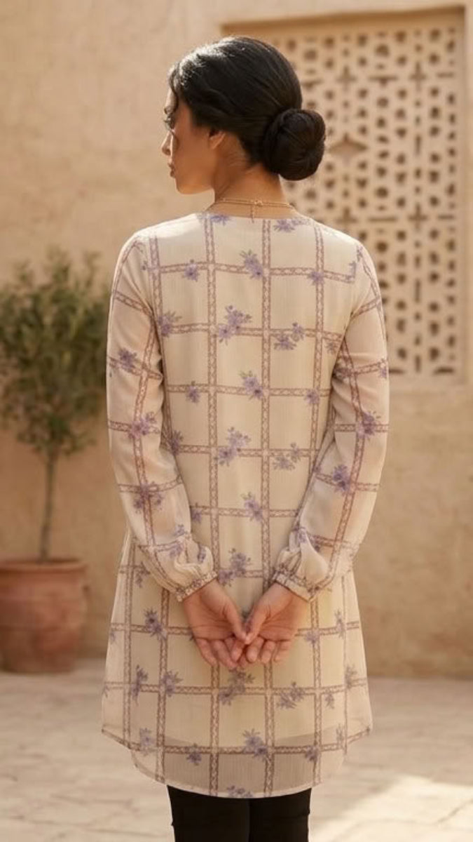 A woman viewed from behind wearing a beige long-sleeve tunic with a purple and green geometric pattern, standing in a sunlit courtyard with potted olive trees and stucco walls.