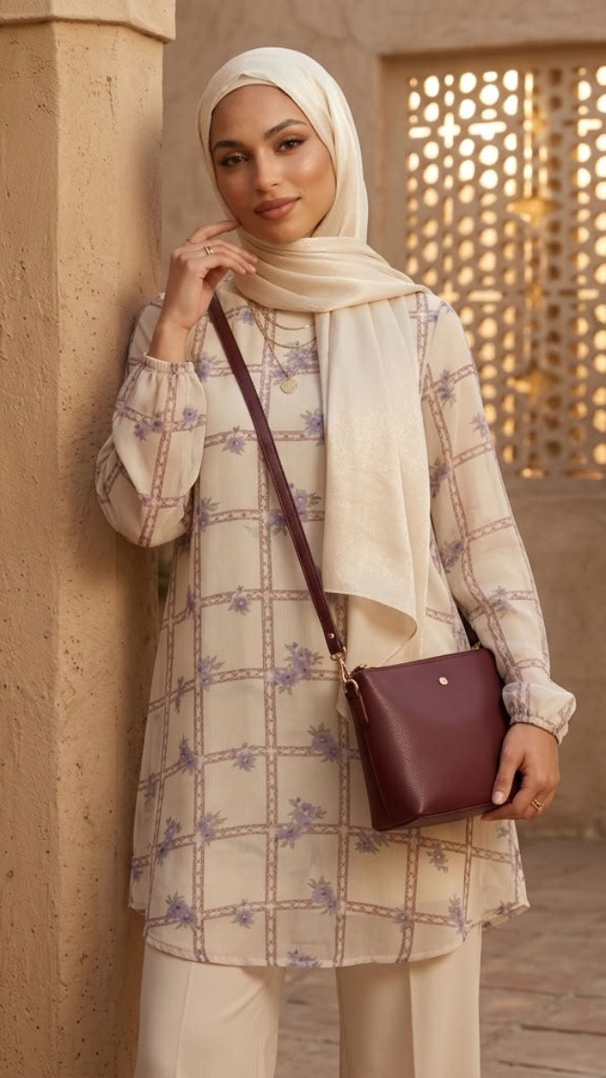 A woman wearing a cream-colored long-sleeve tunic with a purple floral grid pattern, matching cream trousers, and a cream hijab, carrying a dark burgundy crossbody bag. She is posing in a warm, sunlit courtyard with beige stucco walls and columns.