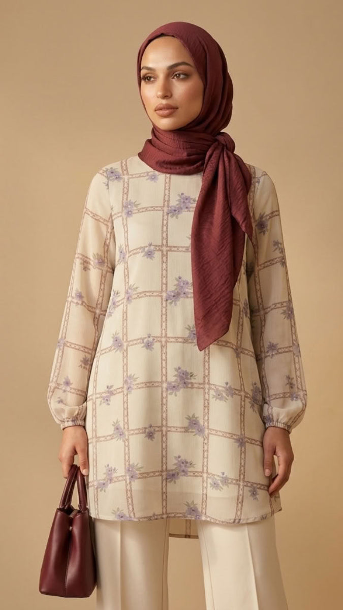 A woman wearing a cream-colored long-sleeve tunic with a purple floral grid pattern, paired with cream trousers and a deep burgundy hijab, holding a matching burgundy handbag against a warm beige background.