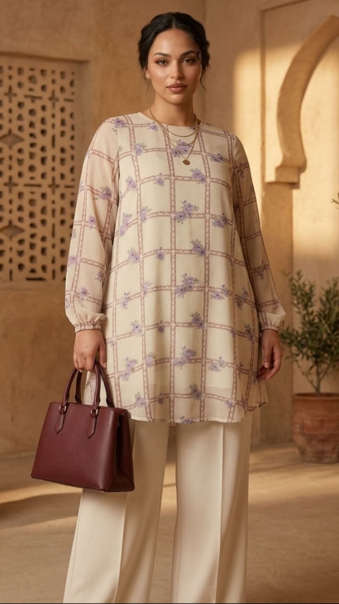 A woman models a beige long-sleeve tunic with a purple floral grid pattern, paired with cream wide-leg trousers and holding a dark burgundy handbag, set against a warm, sunlit desert architectural background with potted plants.