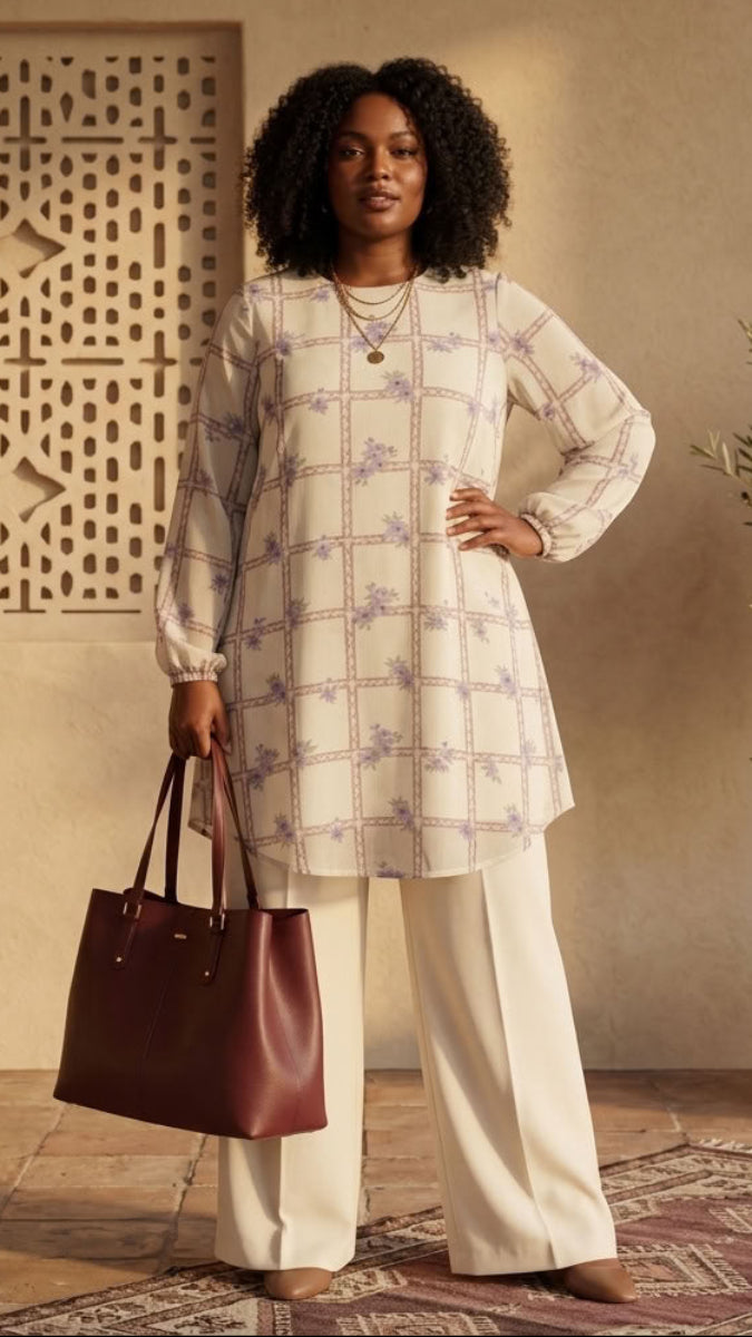 A woman stands in a warm, rustic interior wearing a cream tunic with a light purple and green geometric pattern, paired with cream wide-leg pants and holding a dark brown leather tote bag.