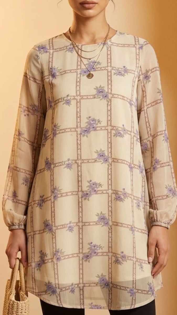 A woman models a long-sleeve beige tunic top with a purple and brown grid pattern and small floral motifs, standing in front of a warm-toned arched doorway.