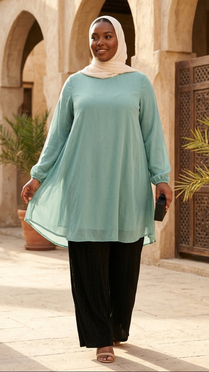 Woman in a light blue dress and black pants standing in a courtyard with arches and plants.
