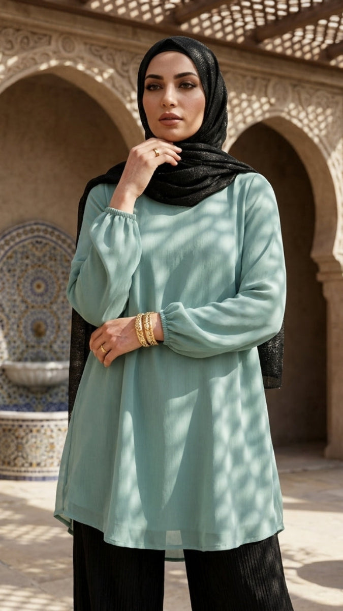 Woman in a light green top and black hijab standing in a sunlit courtyard with arches and plants.