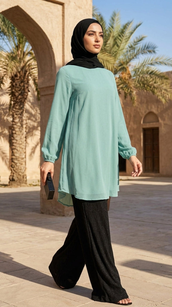 Woman in a light blue top and black hijab standing in a sunlit outdoor setting with palm trees and arches.