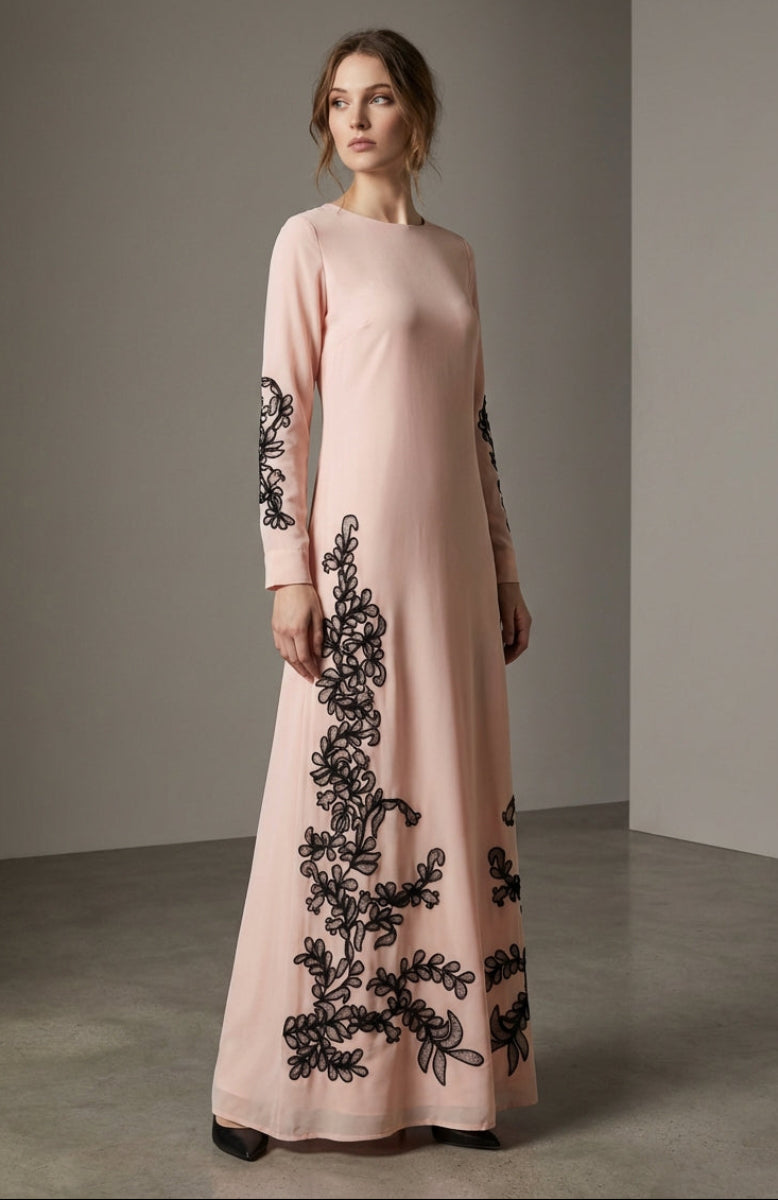 Woman wearing a long, light pink dress with black floral embroidery against a neutral background