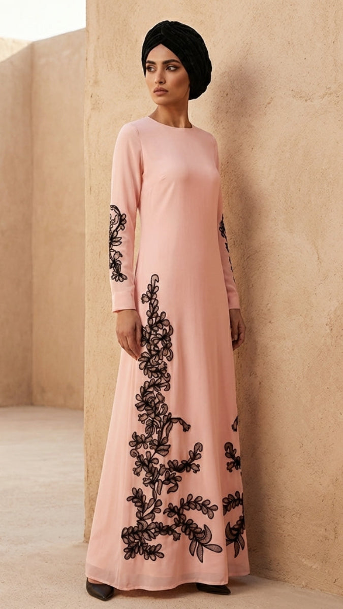 Woman wearing a pink dress with black floral embroidery against a beige wall.