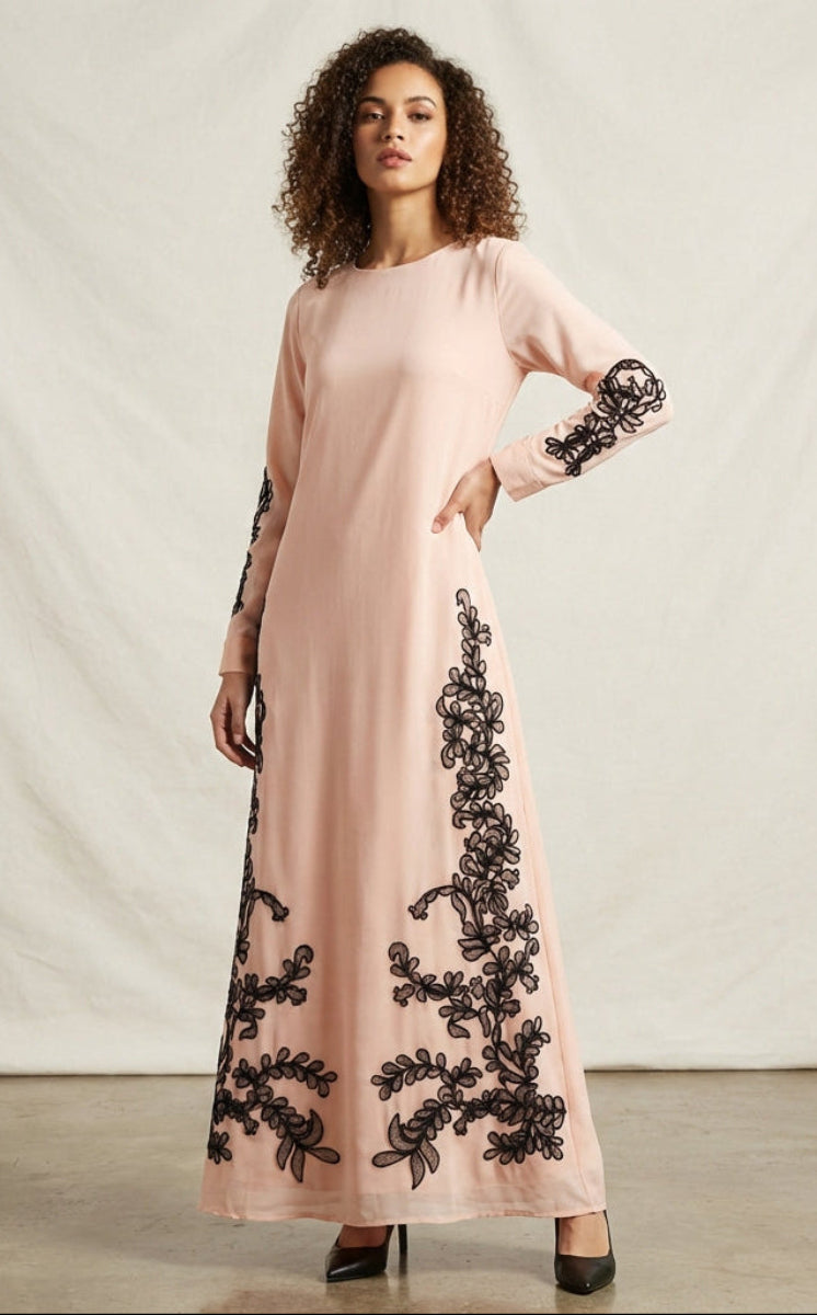 Woman wearing a long pink dress with black floral embroidery against a plain background