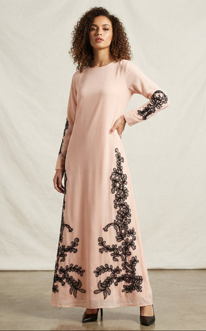 Woman wearing a long pink dress with black floral embroidery against a plain background