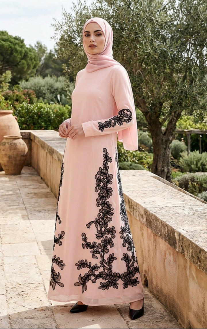 Woman in a pink dress with black embroidery standing outdoors in a garden setting.