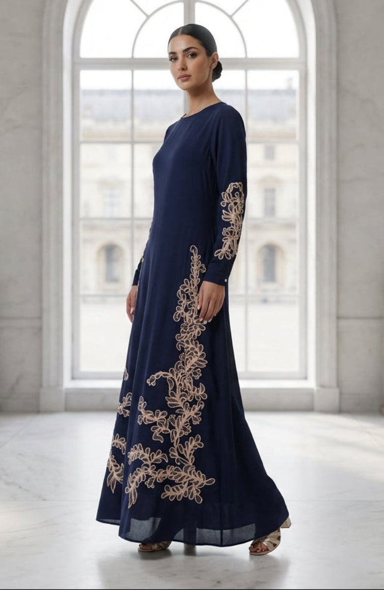 Woman in a navy blue dress with gold embroidery standing in a bright room with large windows.
