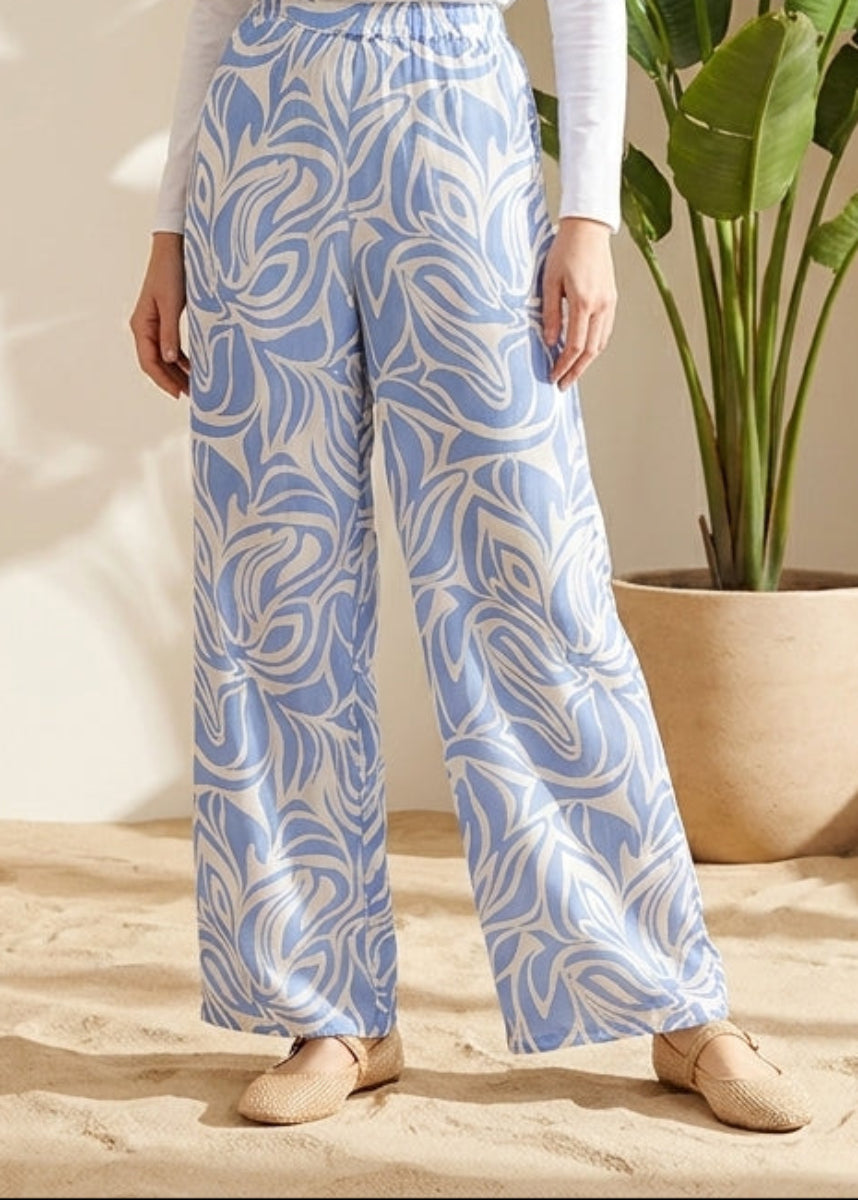 Woman wearing a white top and blue patterned pants standing in a bright room with a plant and woven screen.