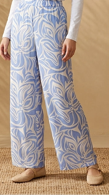 Woman wearing a white top and blue patterned pants standing in a room with a plant and beige wall.