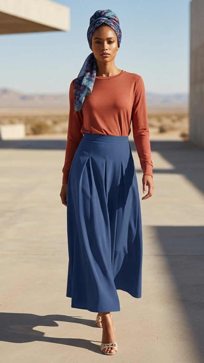 Woman in a blue skirt and brown top standing in a desert-like setting with modern architecture.
