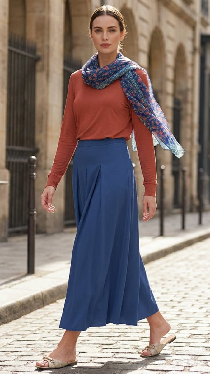 Woman in a blue skirt and red top with a colorful scarf walking on a street.