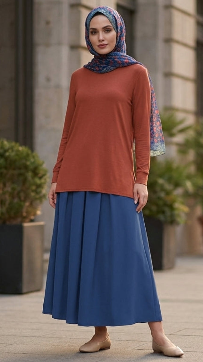 Woman in a red top and blue hijab standing on a city street with plants and buildings in the background.