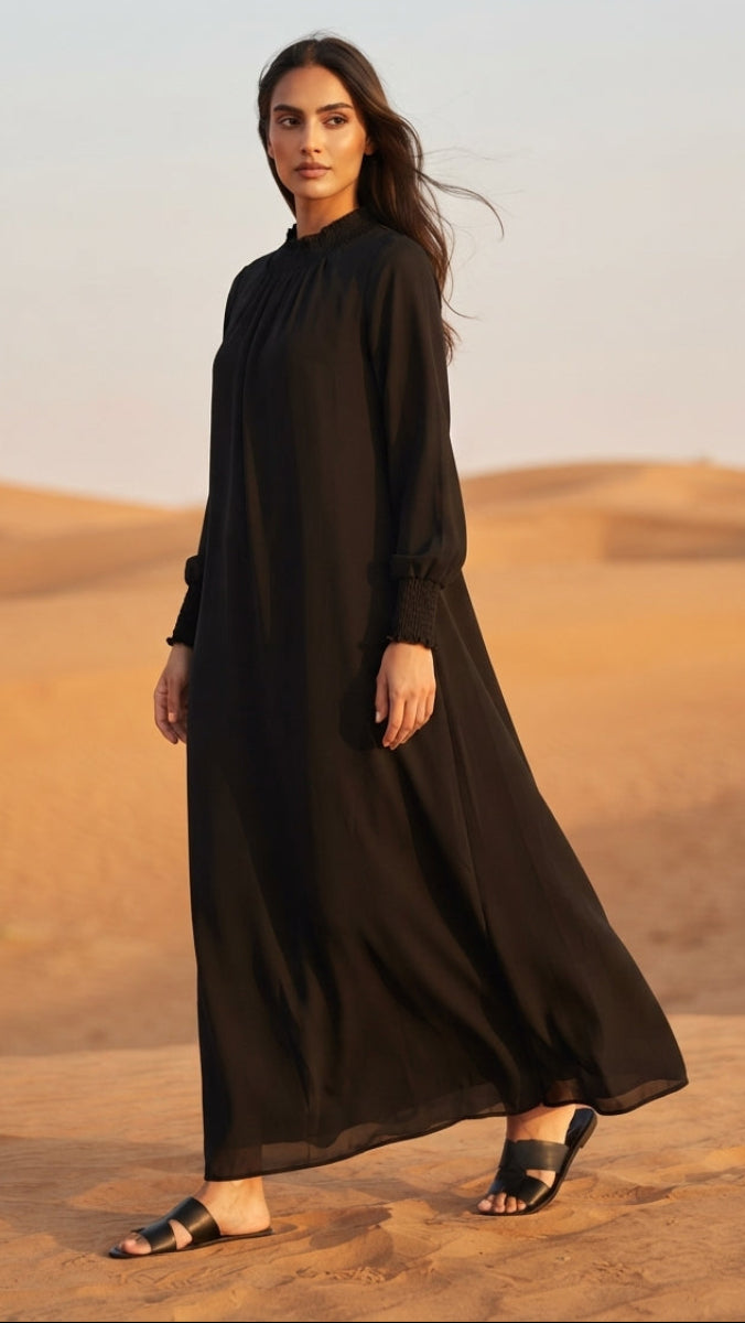 A woman wearing a long black maxi abaya with long sleeves and black slide sandals, standing in a desert landscape with sand dunes under a pale sky.