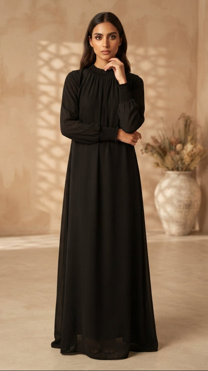 A woman wearing a long-sleeved black maxi dress with a high gathered neckline, standing in a warm beige architectural interior with soft shadows and a decorative vase in the background.