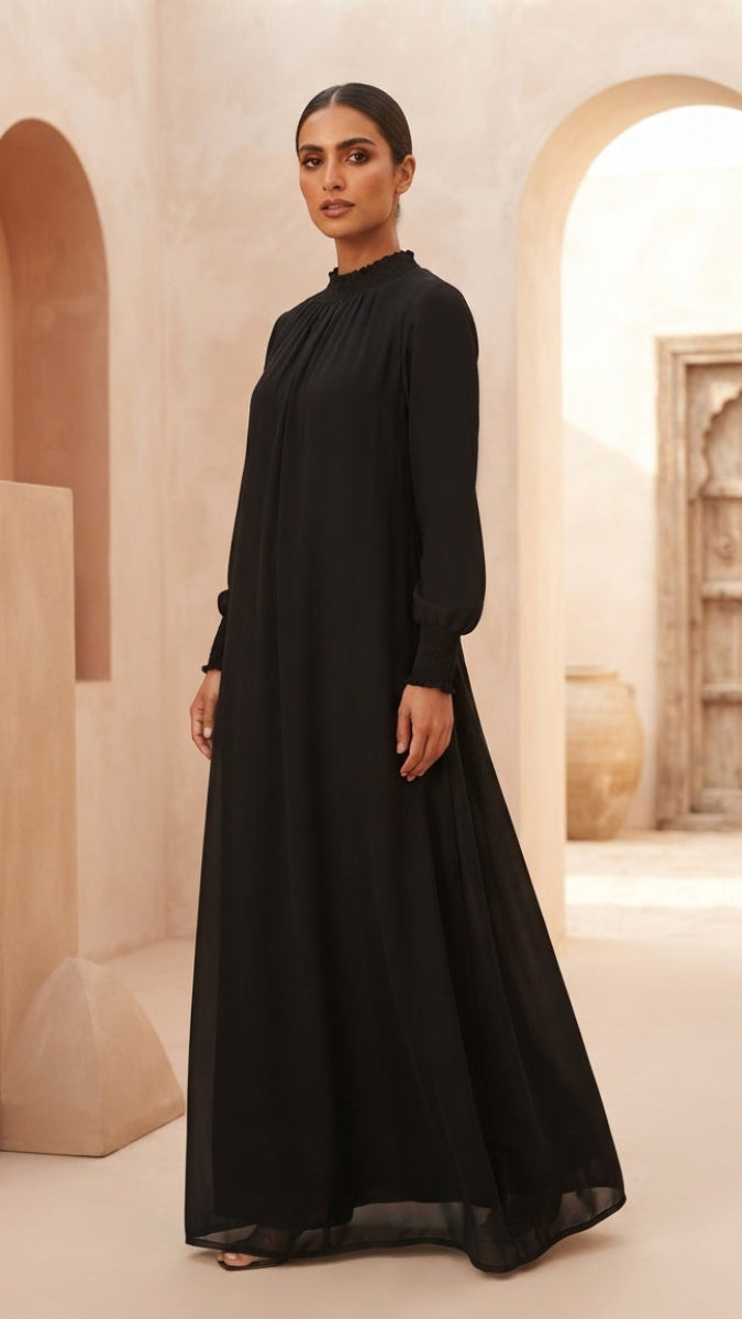 A woman modeling a long-sleeved black maxi dress with a high gathered neckline in a warm, sandy-colored architectural setting with arches.