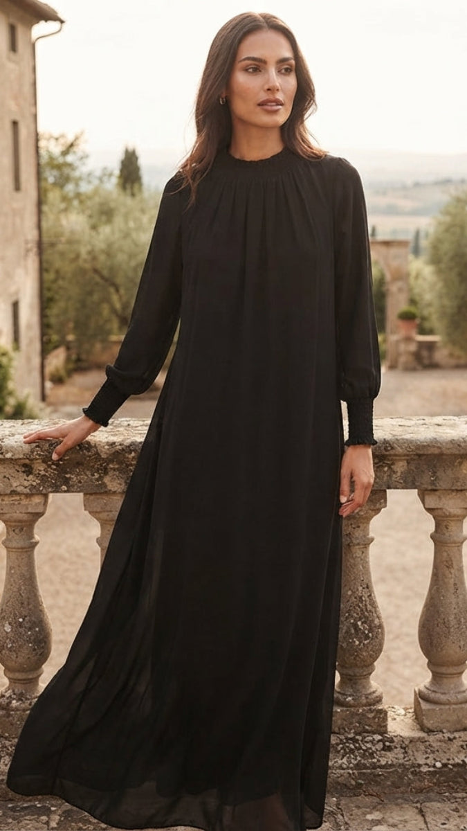 A woman wearing a long black dress stands on a stone terrace outdoors, with a rustic building and olive trees in the background.