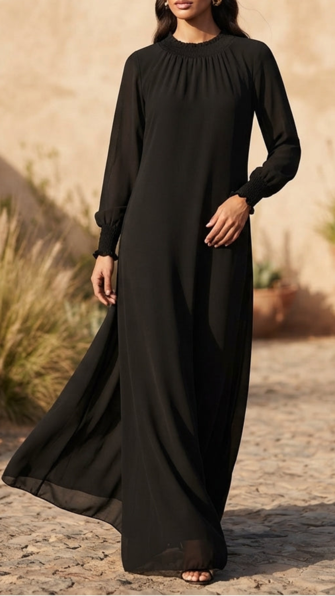 A woman wearing a long black flowing dress with long sleeves, standing outdoors against a warm beige stone wall and cobblestone ground.