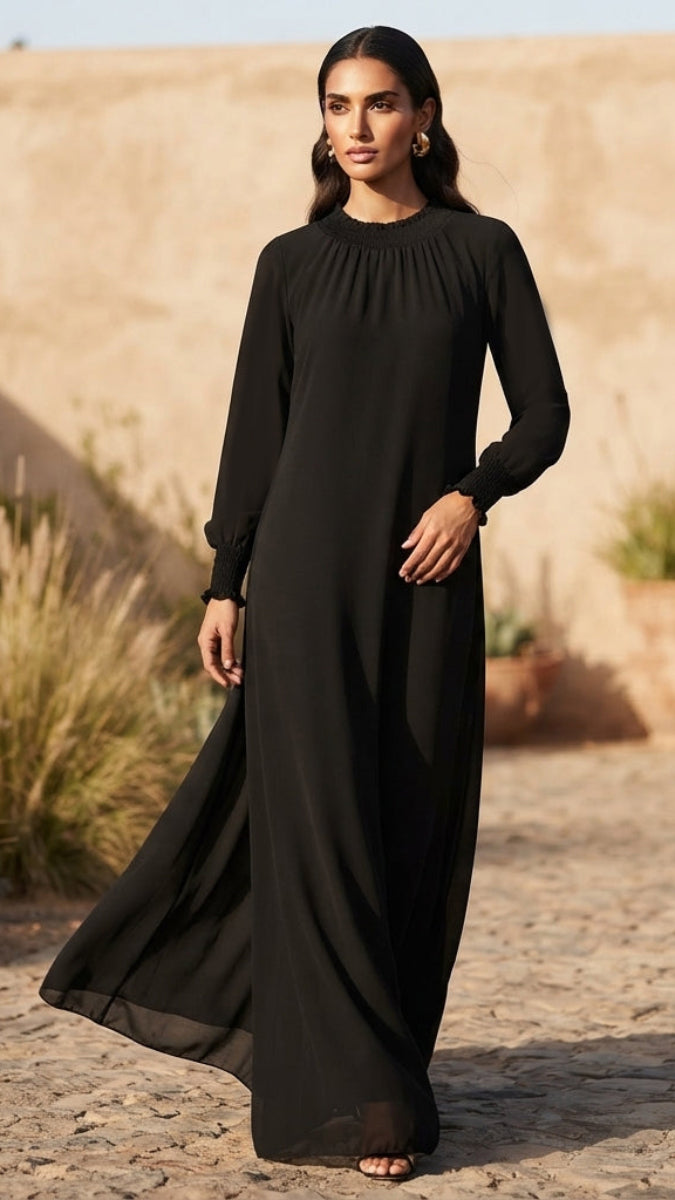 A woman wearing a long-sleeved black maxi dress stands outdoors against a sandy, desert-like background. The dress has a gathered high neckline and a flowing silhouette.