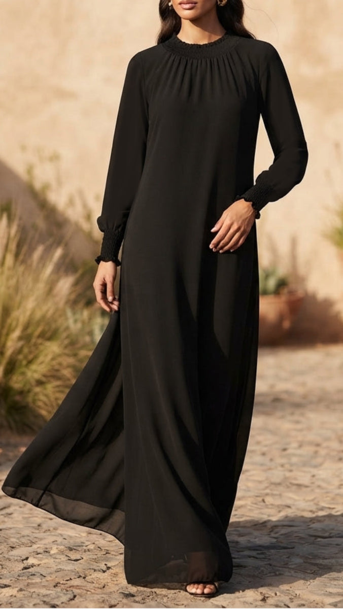 A woman wearing a long-sleeved black maxi dress with a gathered high neckline, standing outdoors on a stone path against a warm beige wall.